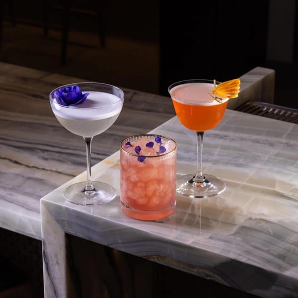 Three colorful cocktails, garnished with flowers and orange peel, on a marble bar top.