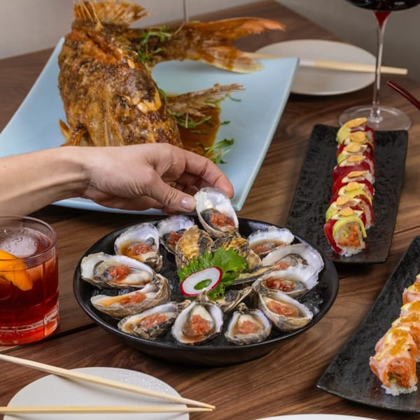 A hand reaching for an oyster, with a whole fried fish, sushi rolls, and drinks on a wooden table.