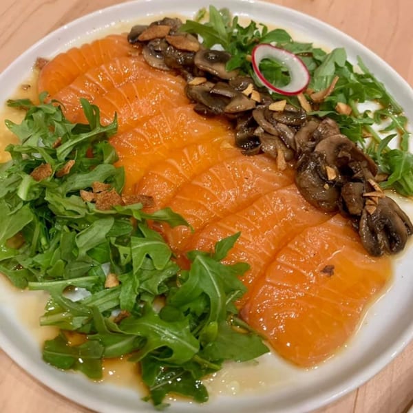 Yume Sushi Bar's salmon carpaccio with arugula, mushrooms, and a radish slice on a white plate.