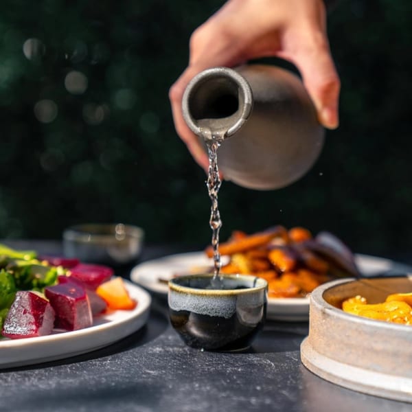 A hand pours sake from a ceramic bottle into a small cup, with sushi dishes in the background.