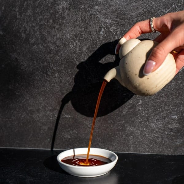 A hand pours dark soy sauce from a ceramic pot into a small white dish on a dark surface.